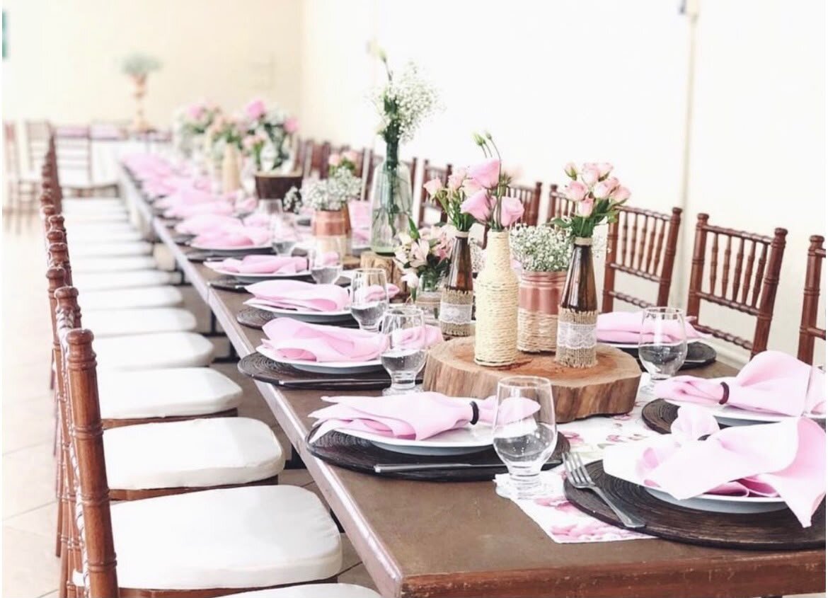 Detalhe da mesa posta da Casa 1: pratos, taças, guardanapos rosa e arranjos de flores.