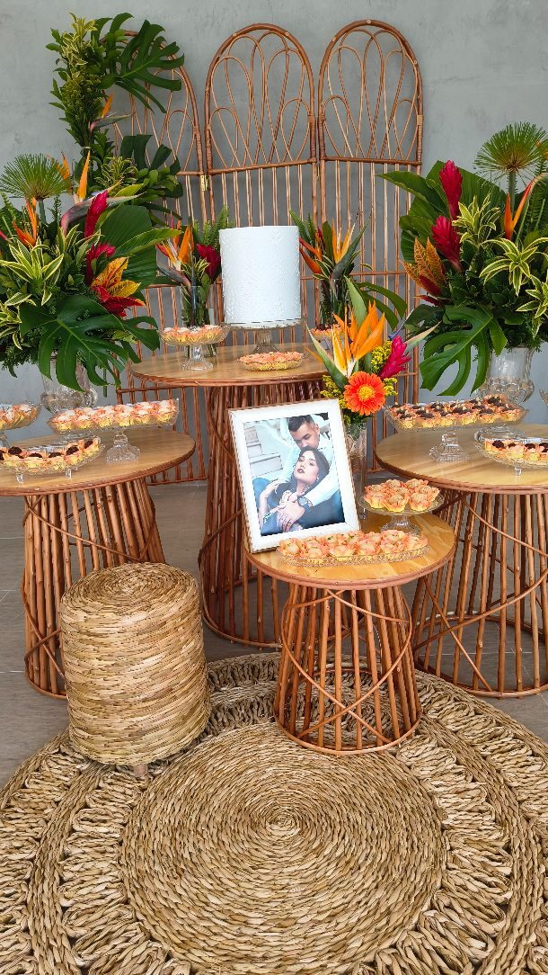 Mesa de doces montada com arranjos tropicais de folhas e flores alaranjadas.